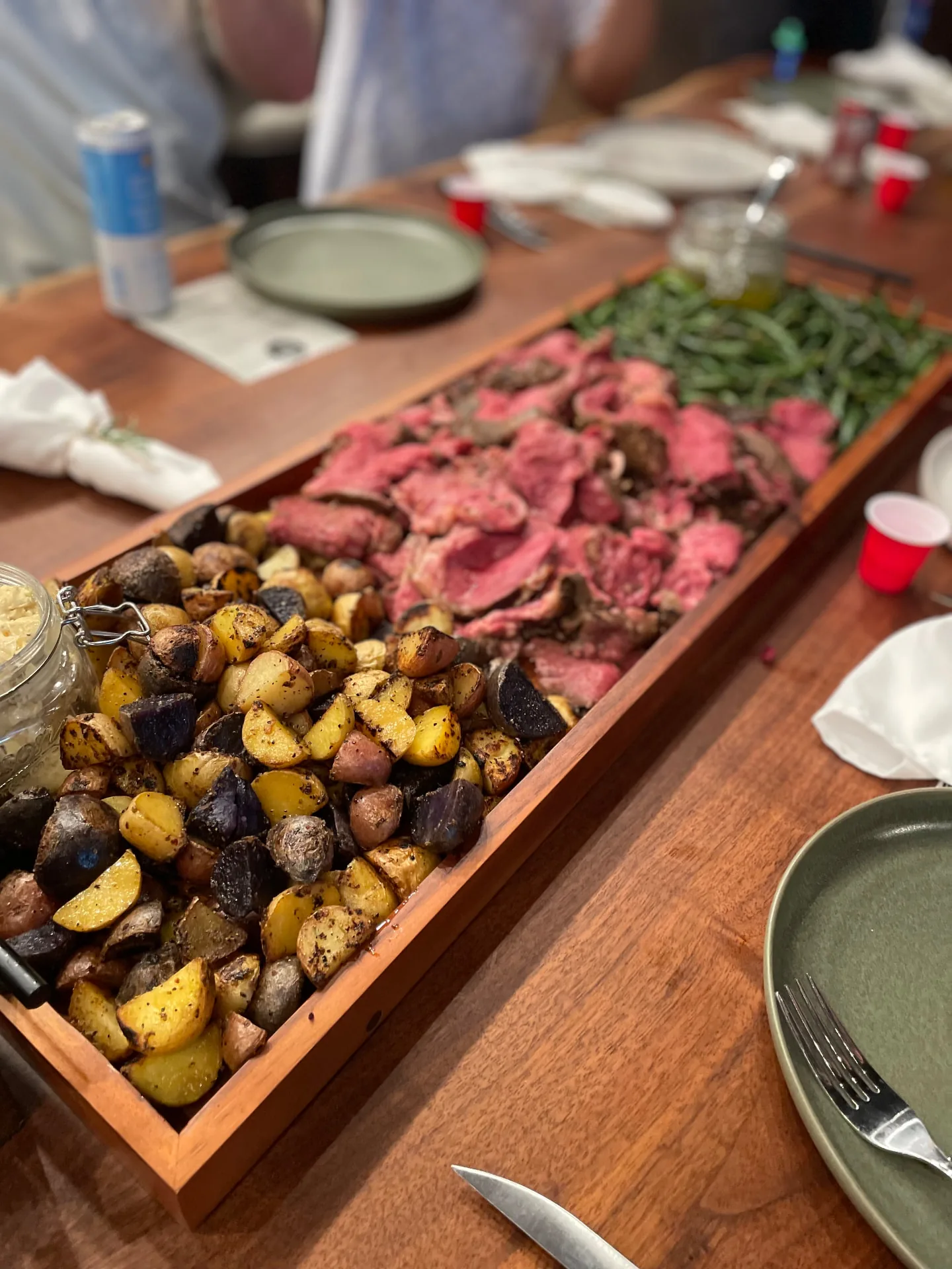 Family-style serving board of sliced roast beef, roasted potatoes and green beans set down a long table